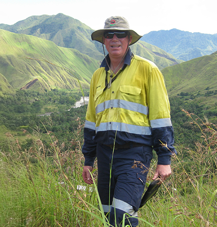 David wearing high-visibility yellow and navy workwear standing in tall grass on a green hillside, with rolling mountains and a valley in the background.