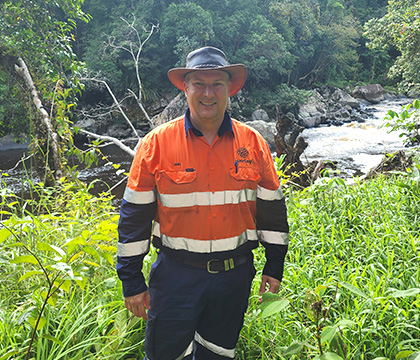 Iain wearing Worley high-visibility orange and navy blue safety workwear and a wide-brimmed hat, standing outdoors in lush green foliage near a river in a forested area.