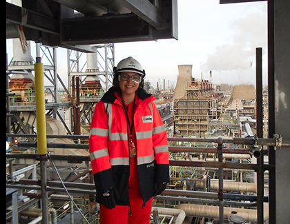Katie in high-visibility safety gear and a helmet standing on an industrial platform with a large refinery and smoke in the background.
