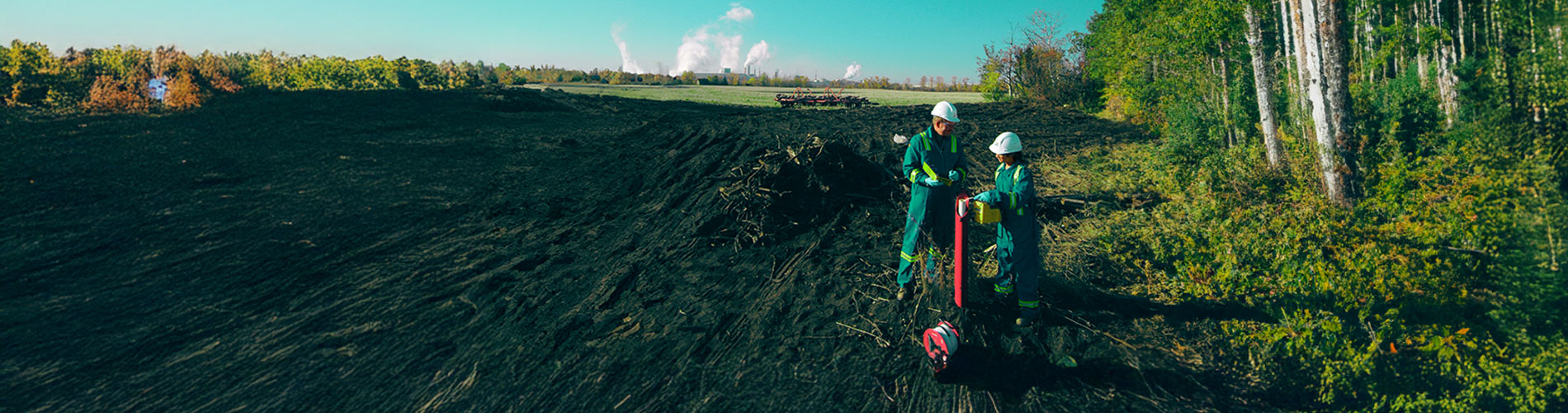 Two people wearing PPE working on soil with machinery next to a forest.