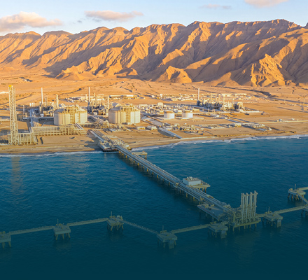 Coastal industrial processing facility with storage tanks, marine loading jetty and mountainous desert landscape.