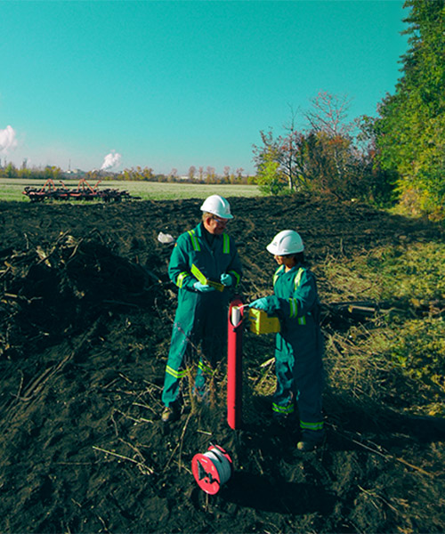 Two people wearing PPE working on soil with machinery next to a forest.