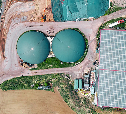 Overhead view of a biogas plant with storage tanks and a solar-roofed building in the middle of crop fields.