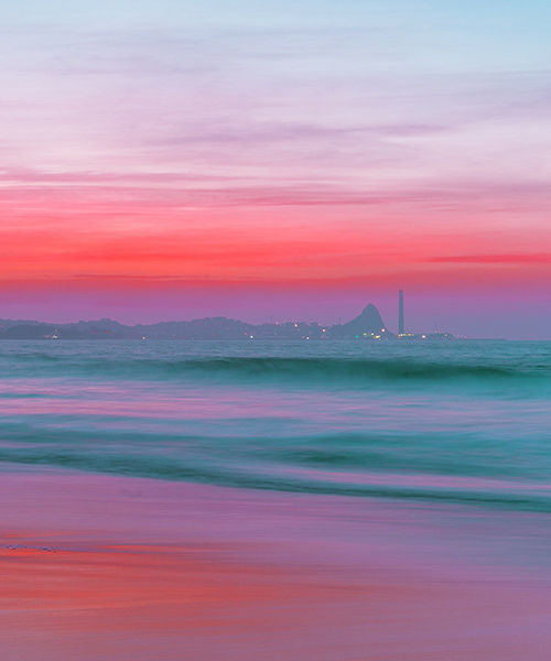 Looking over a body of water to a refinery and a pink sky.