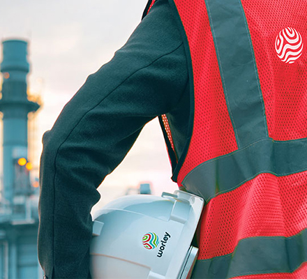 Person wearing PPE holding a Worley safety helmet under his arm in front of a refinery.