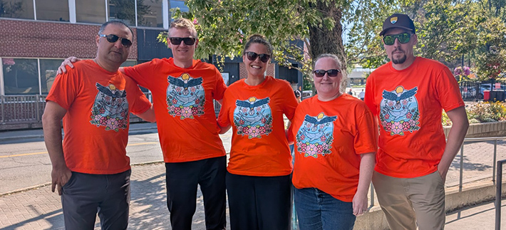Five people standing together wearing bright orange tshirts with Indigenous artwork on the front.