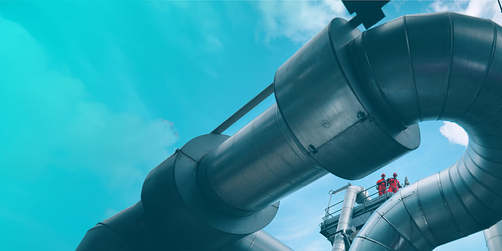 Looking upward at two people standing on a platform, past large steel pipes at a plant,