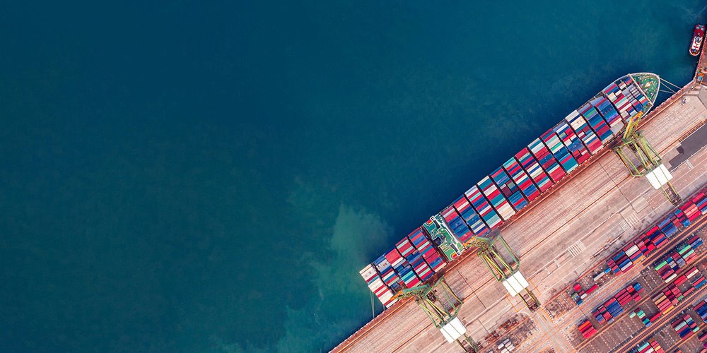 Aerial view of a large container ship docked at a busy port, surrounded by cranes and other vessels.