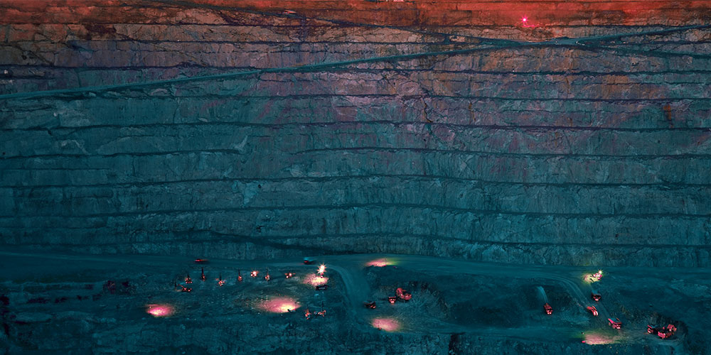 Looking into an open mine with several mining vehicles with lights on.