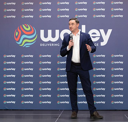 Patrick standing on stage speaking into a microphone in front of a Worley branded wall.