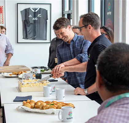 Tom smiling and cutting a cake with a colleague in an office kitchen.
