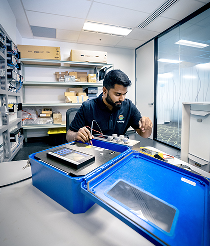 Worley technician testing electronic components in a lab with tools, blue case, and shelves of equipment.