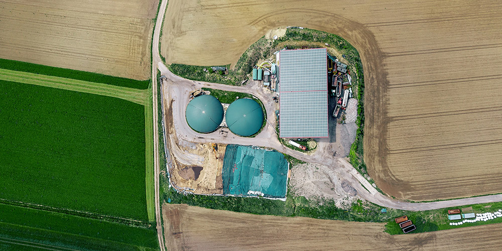 Overhead view of a biogas plant with storage tanks and a solar-roofed building in the middle of crop fields.