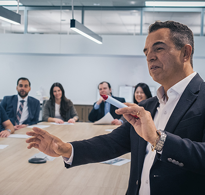 Four people sitting at the end of a boardroom table listening to a man at the front pointing to something.