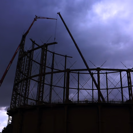 Steel framework of a gasometer with cranes positioned around it against a dark, cloudy sky.