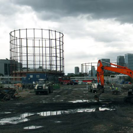 Construction site with an excavator and trucks in muddy ground, beside a steel gasometer frame.