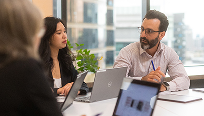 Three people sitting at a desk together talking with their laptops in front of them.