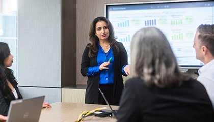 Three people sitting around a table listening to a woman presenting in front of a TV screen.